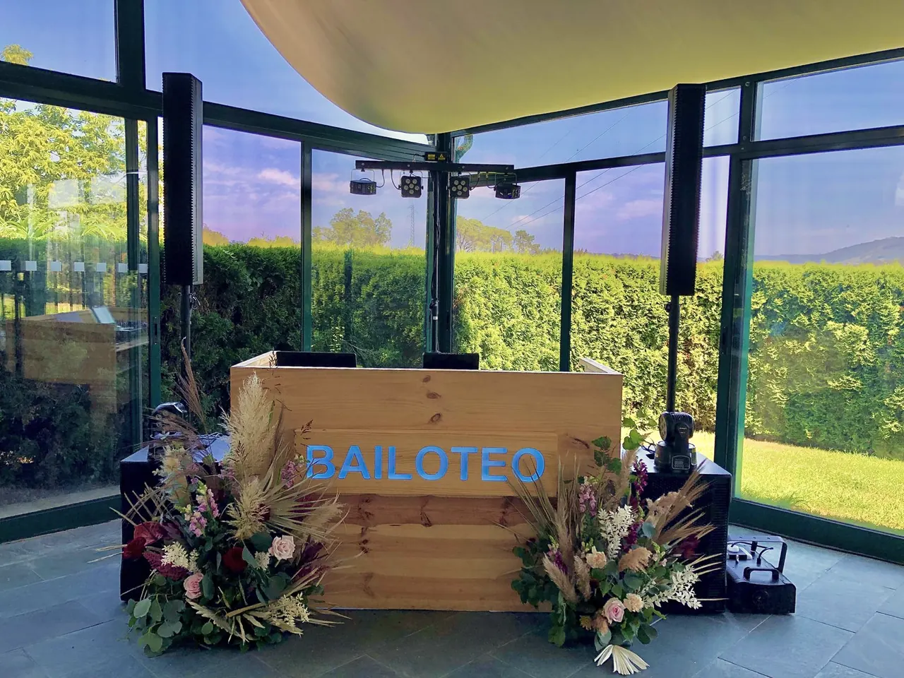 Cabina DJ de madera natural con letras «BAILOTEO» y arreglos florales en un pabellón acristalado