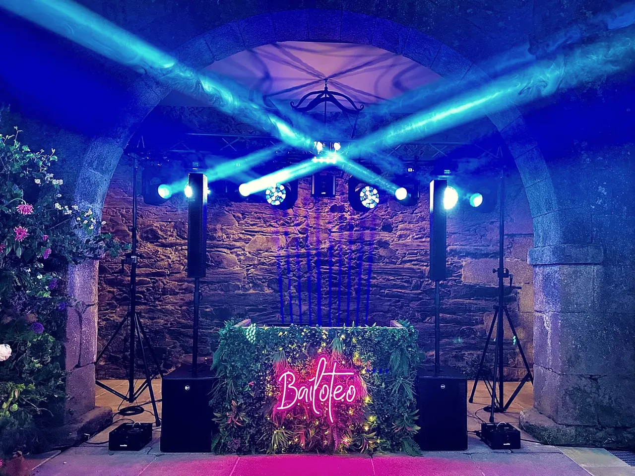 Cabina DJ vegetal con neón rosa «Bailoteo» bajo un arco de piedra, con cabezas móviles en azul