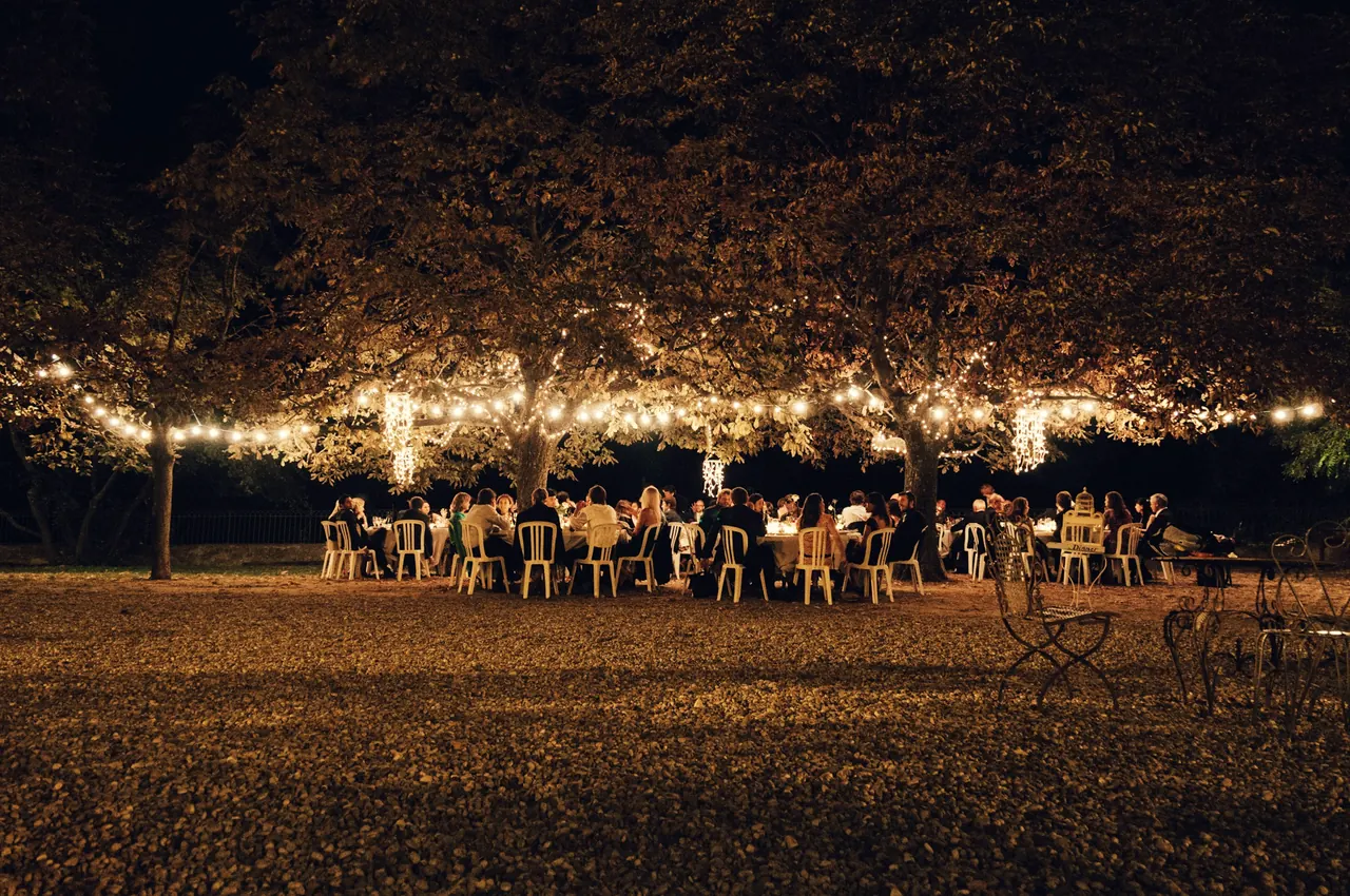 Cena de boda al aire libre, de noche, bajo árboles con guirnaldas de luces